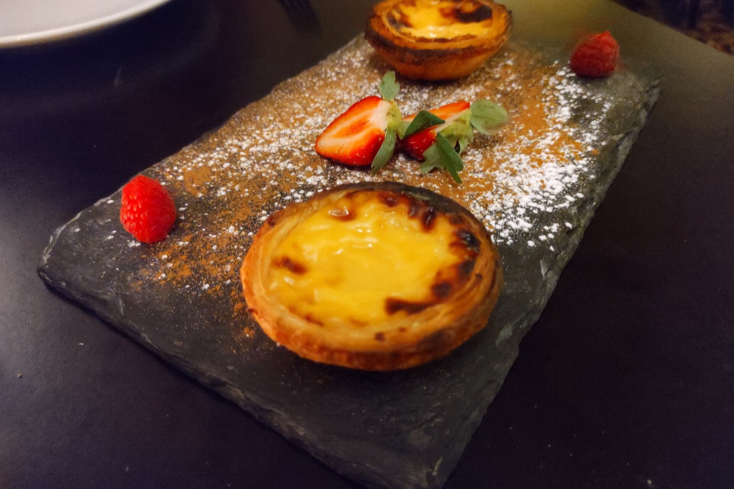 Two pastéis de nata on a slate board with berries and icing sugar