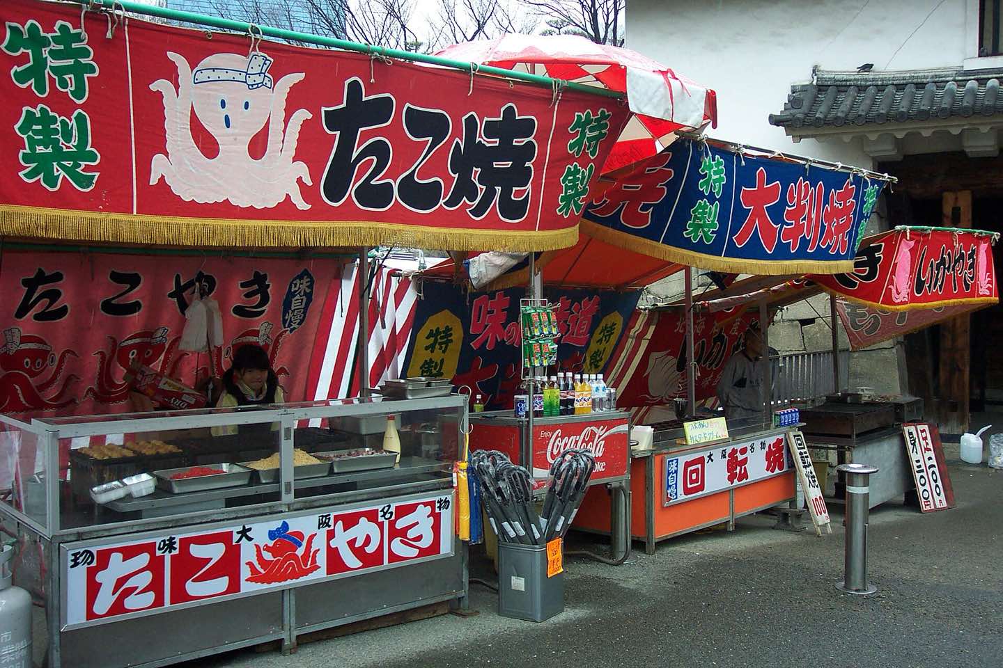 Red-bannered takoyaki stalls with griddles and display cases in Osaka