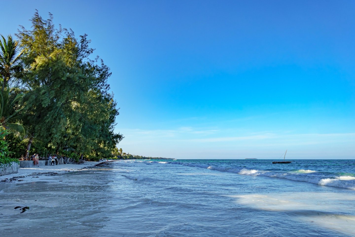 Waves on a white-sand beach lined with trees at Nungwi, with a dhow offshore
